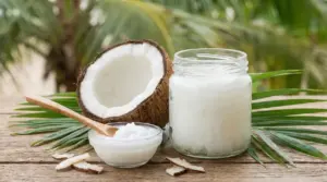 Barattolo e ciotola di olio di cocco con mezzo cocco su tavolo di legno, con foglie di palma sullo sfondo