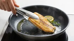 Filetto di pesce in padella antiaderente, girato con pinze da cucina, con limone e rosmarino